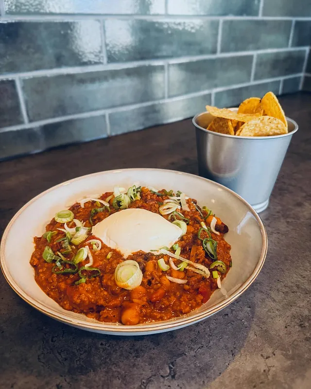 Skål med chili con carne toppet med rømme og vårløk, servert med nachos i metallbeger.